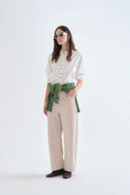 Load image into Gallery viewer, A woman modeling a white button-up shirt with colorful buttons, beige wide-leg trousers, and an olive green tied shirt-waist, styled with green-tinted sunglasses and dark brown loafers against a plain white studio background.