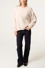 Load image into Gallery viewer, A woman modeling an oversized cream-colored knit sweater paired with dark blue jeans and black shoes, standing against a plain light background.